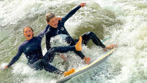 Zwei Jugendliche surfen auf einer Welle, beide in Neoprenanzügen, lachend und voller Freude.