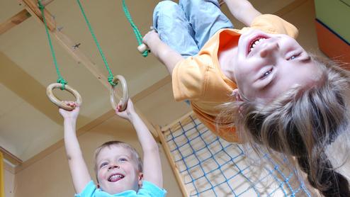 Drei Kinder spielen in einem Raum: Ein Junge hängt an Ringen, während zwei andere lachen und zuschauen.